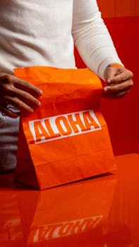 Close-up of hands holding a bright orange 'ALOHA' paper bag on an orange surface. Indoor setting.