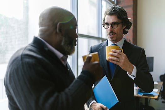Two diverse professionals in corporate attire having a casual conversation over coffee indoors.