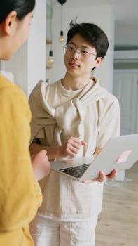 Two young professionals engaging in a discussion, holding a laptop in a modern office setting.